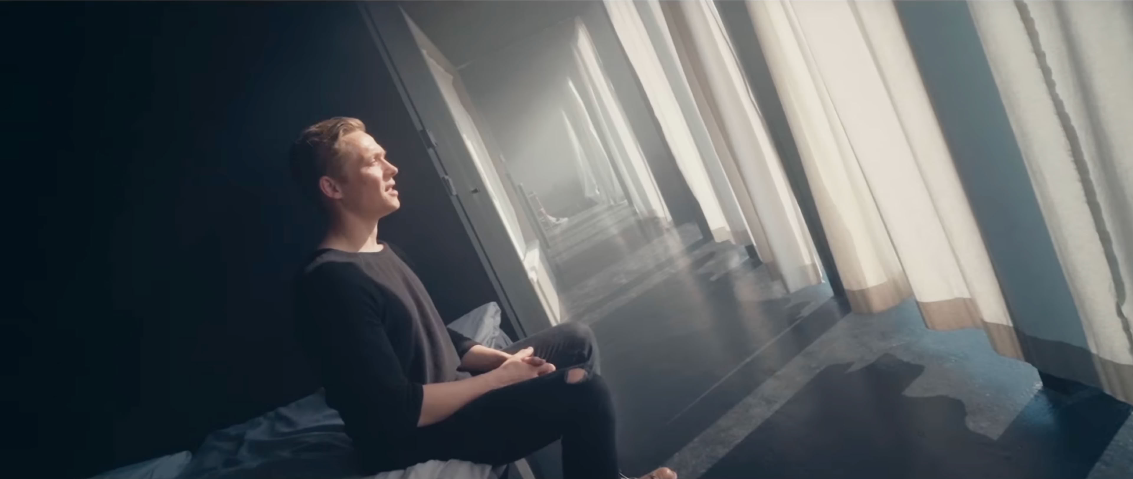 Eine Person sitzt auf einem Bett und schaut aus einem großen Fenster, durch das helles Licht scheint.