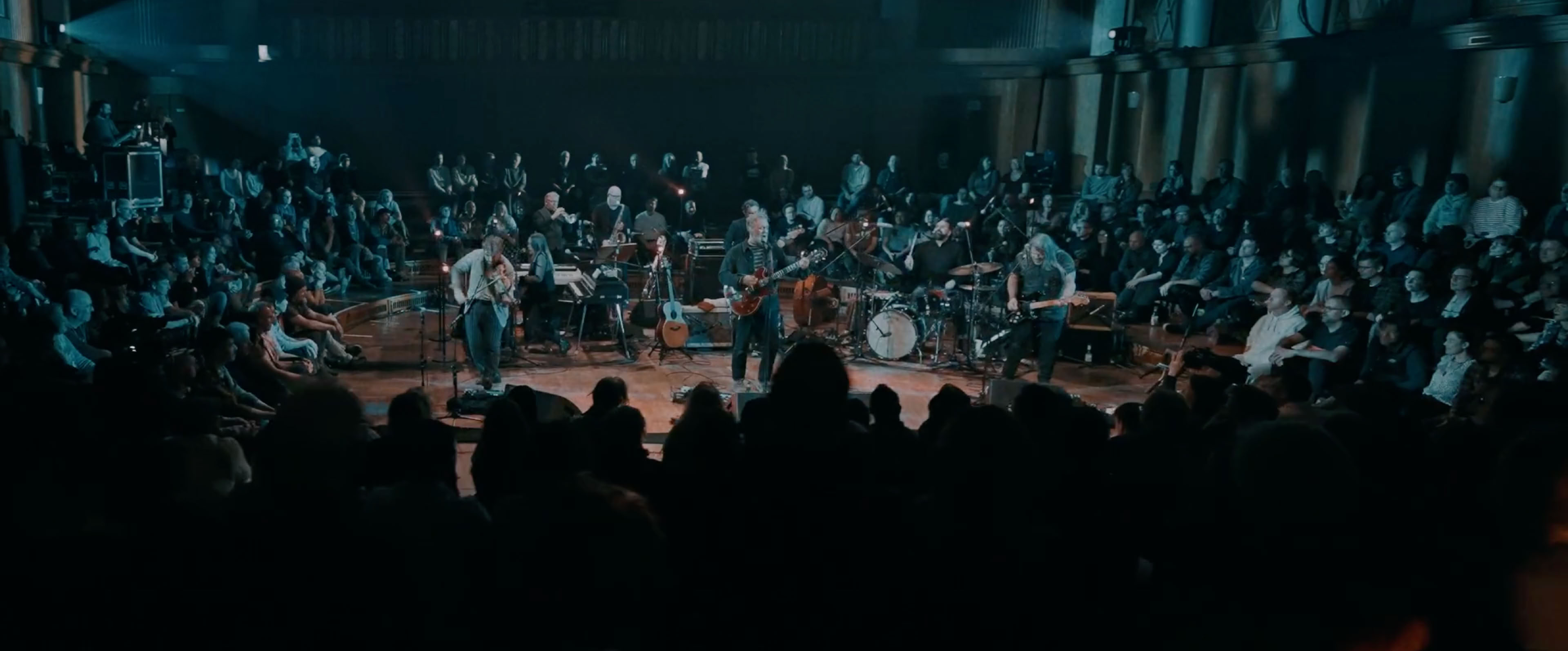 Eine große Menschenmenge sitzt in einem Saal, während eine Band auf einer Bühne in der Mitte spielt.