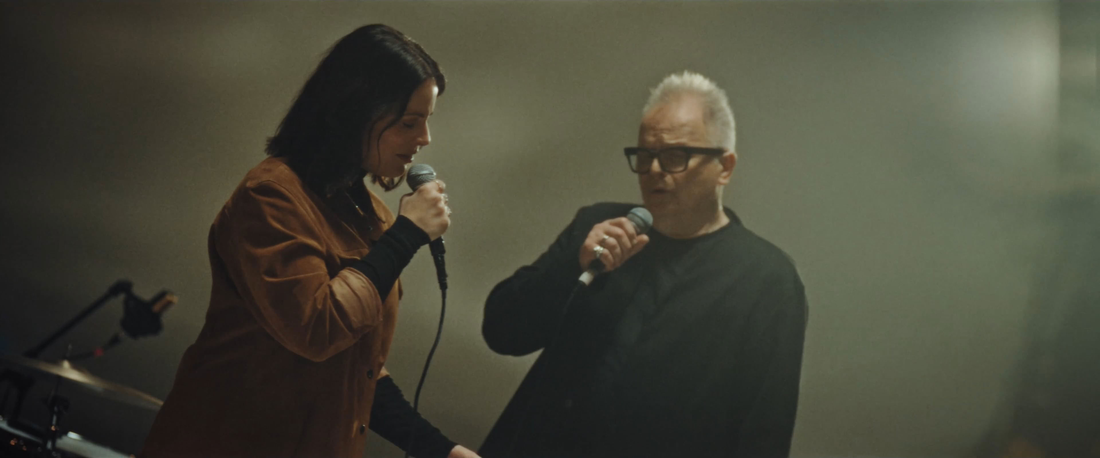 Zwei Personen singen in Mikrofone, eine trägt eine braune Jacke, die andere eine schwarze Brille.